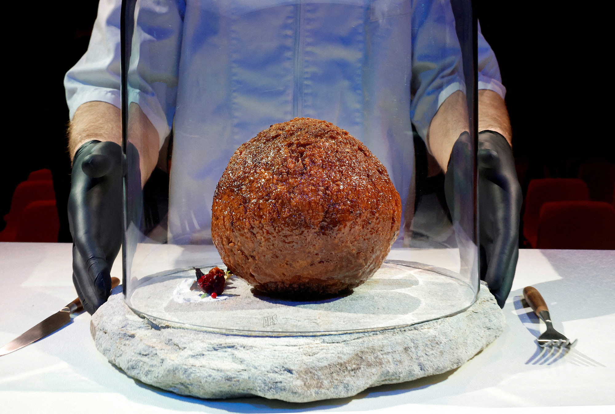 an enormous meatball under a glass container being held by a person in black gloves