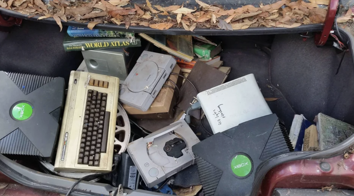 The trunk of a car covered in leaves is popped to reveal a pile of old tech that includes a typewriter, and two original XBox and PlayStation video game consoles