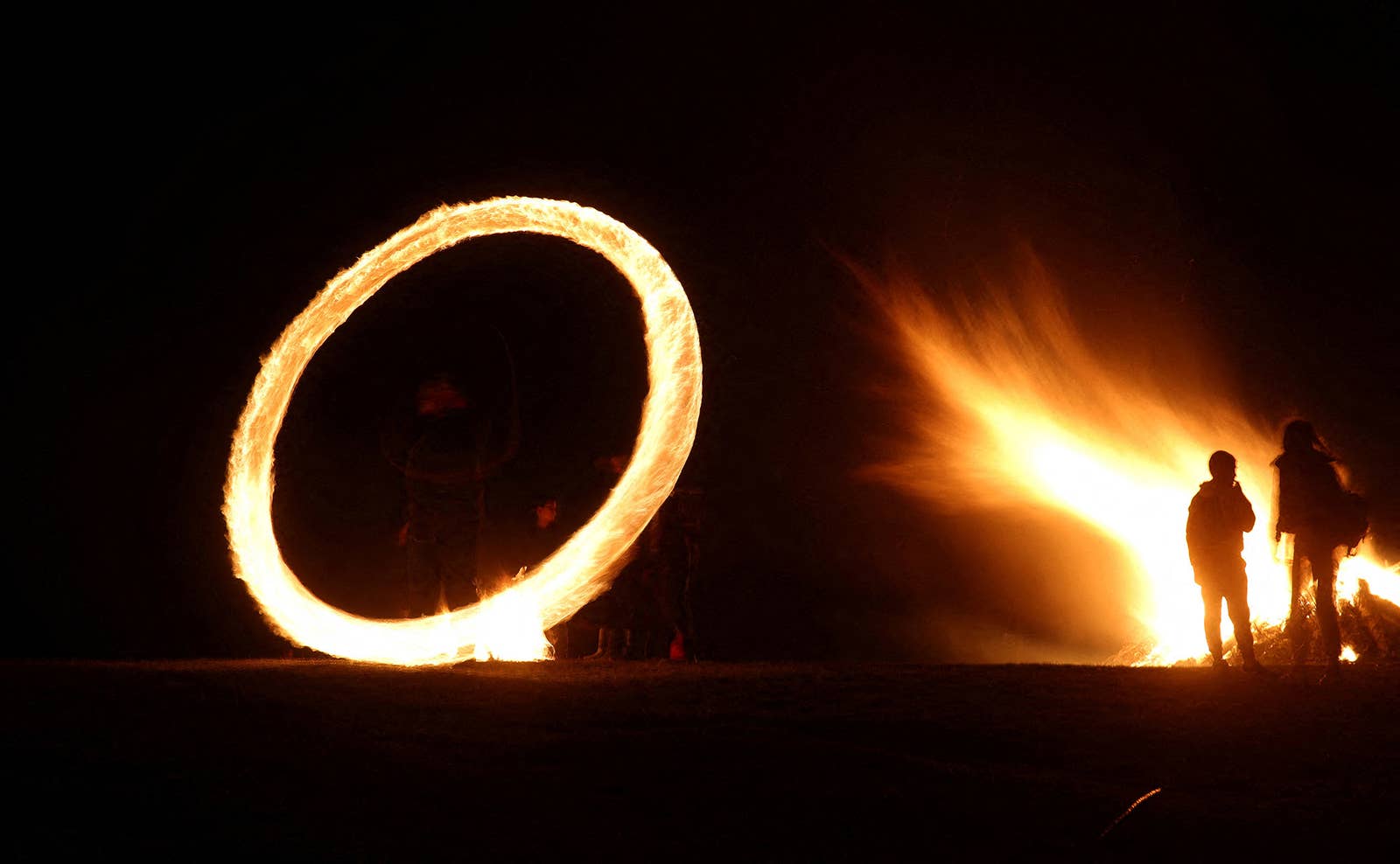 A long-exposure photo showing a ring of fire being spun around someone standing, while two figures are silhouetted by a bonfire to the right in the frame