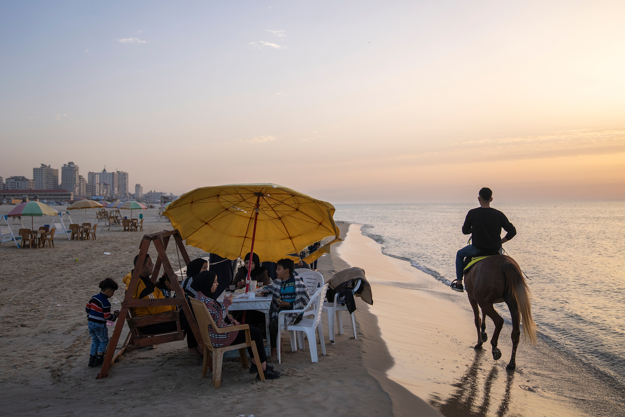 A person rides a horse alongside a shore next to a family gathered around a plastic table and enjoying a meal, seated in chairs and a swinging bench under a big yellow umbrella