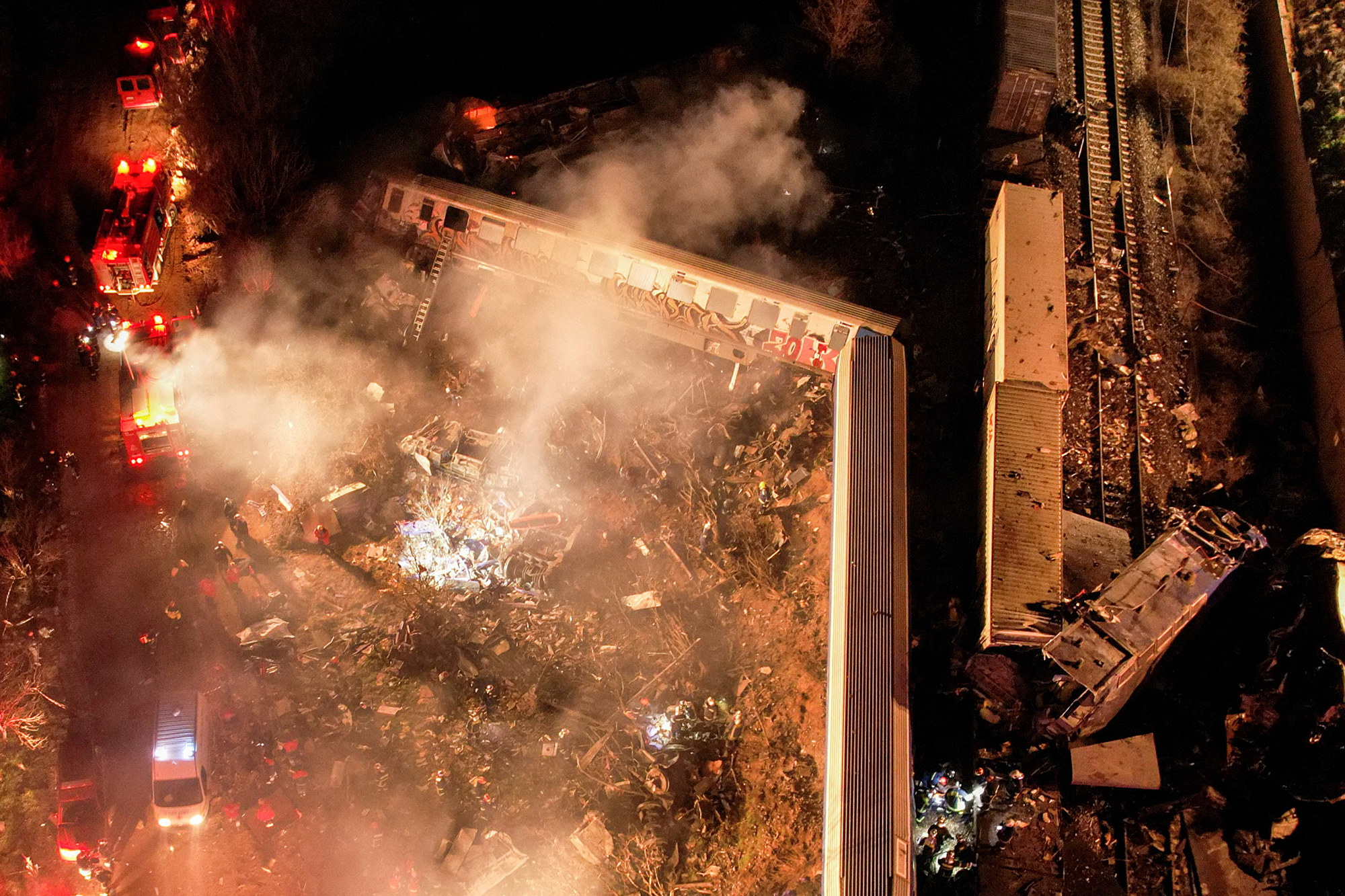 An aerial view at night of derailed freight and passenger train cars, amid debris, smoke, emergency vehicles, and fire trucks with their lights on