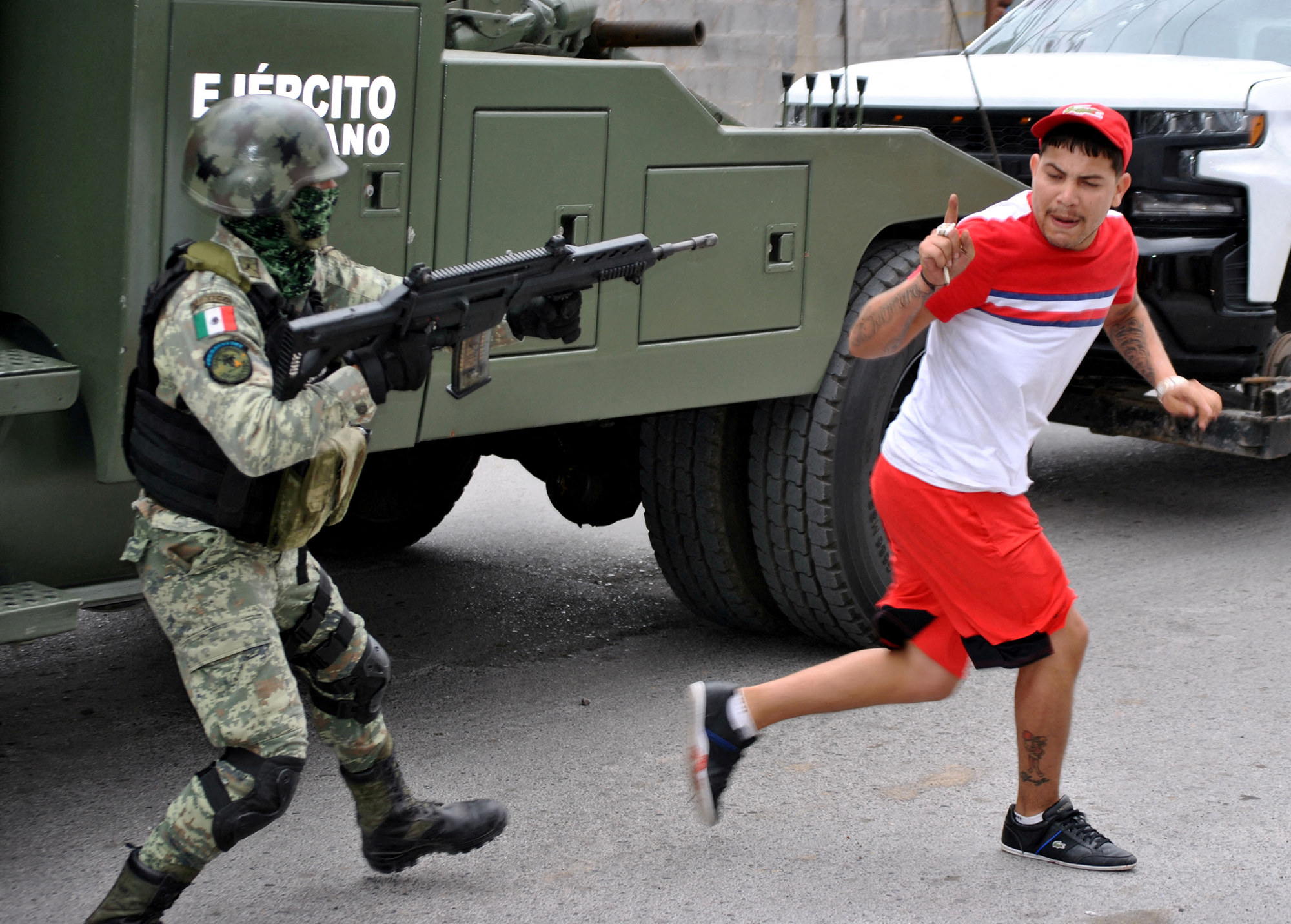 A person wearing red shorts, t-shirt and a baseball cap holds up a finger and runs away from a man holding an automatic gun and is dressed in military garb with boots, camouflage outerwear and helmet in front of a military truck