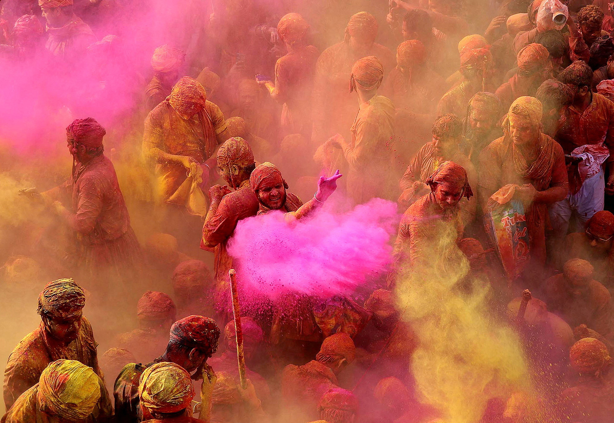 During a Holi celebration, a hazy overhead view of a crowd of people covered in colorful dust amid clouds of yellow and pink powder being tossed at one another