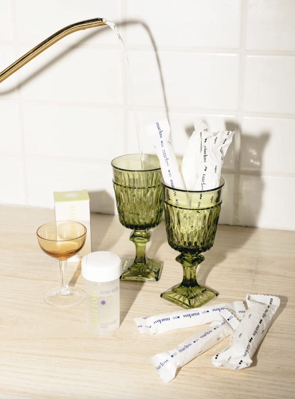 several tampons and a bottle of lube artfully arranged in glassware; a person pours liquid into one of the glasses
