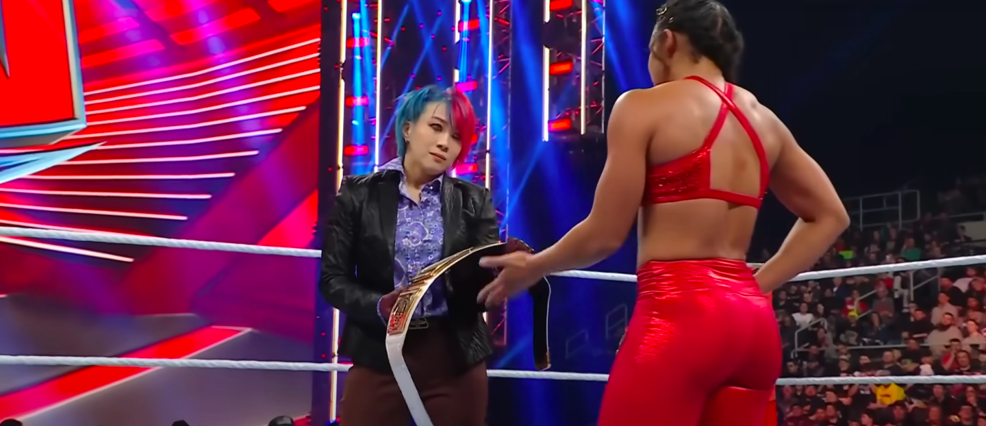 A woman holds out her hand, asking another woman to hand her a title belt
