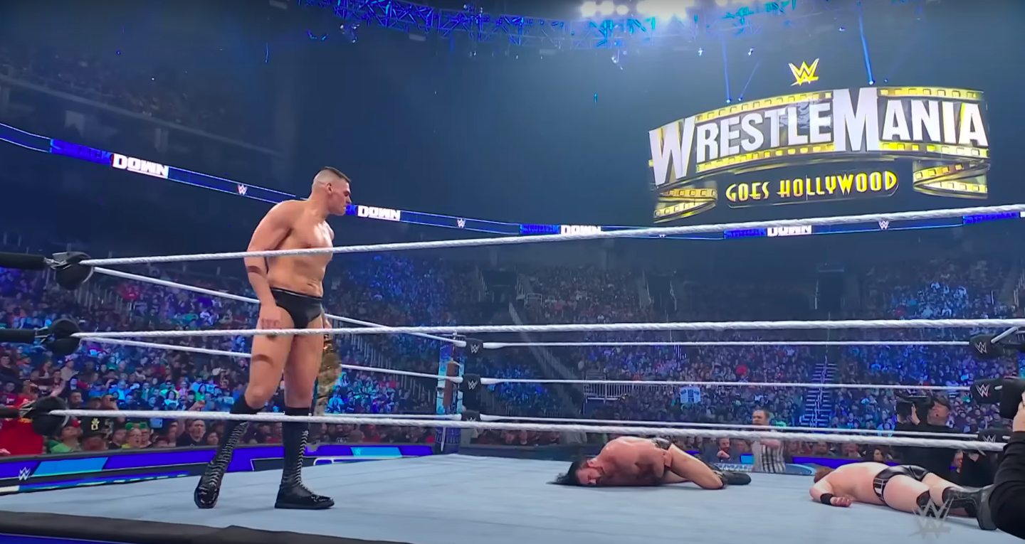 A man stands over the bodies of two other men with the WrestleMania sign in the background