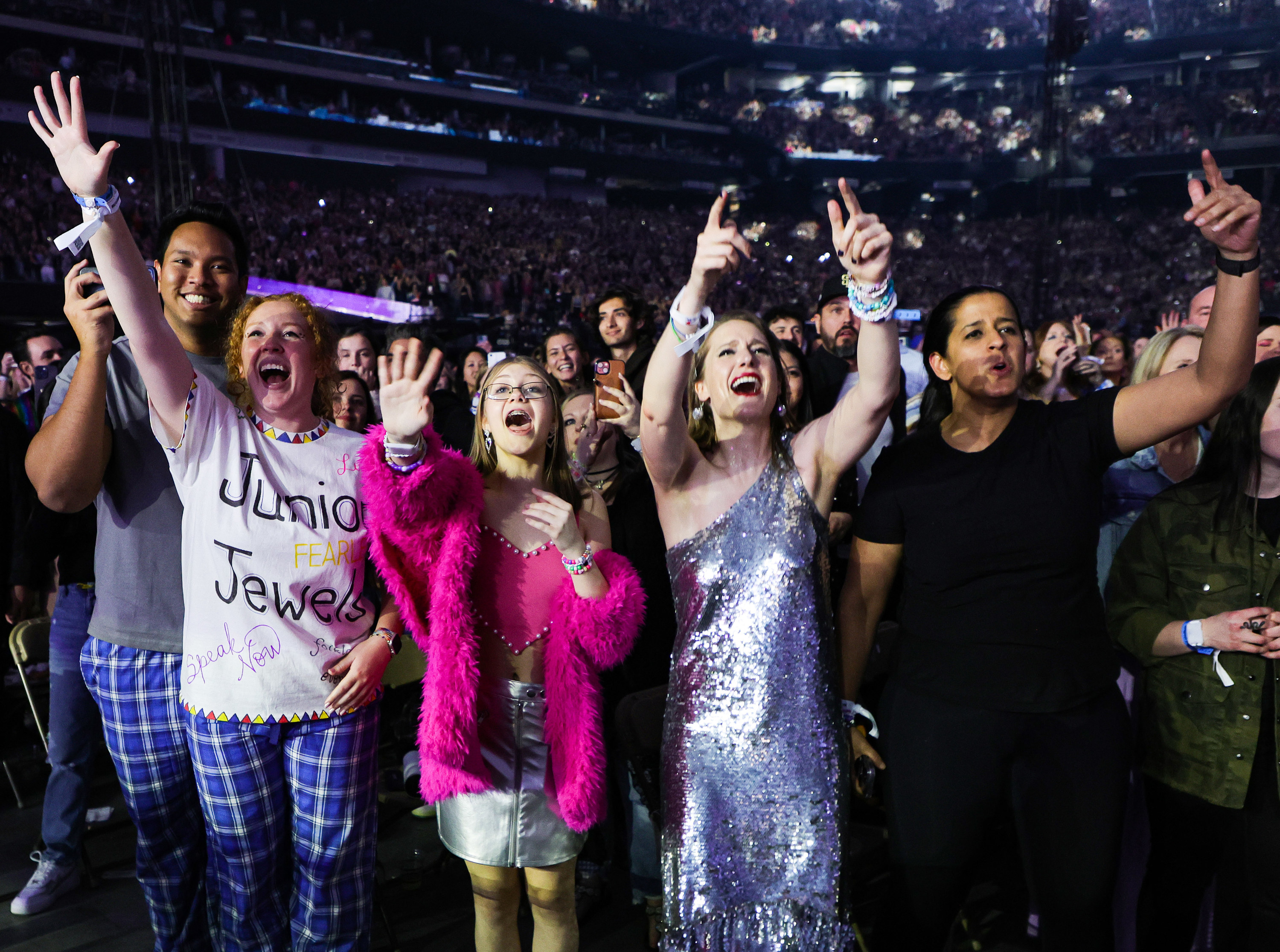 Fans at a concert stand side by side in a packed arena, arms up, all singing along joyfully