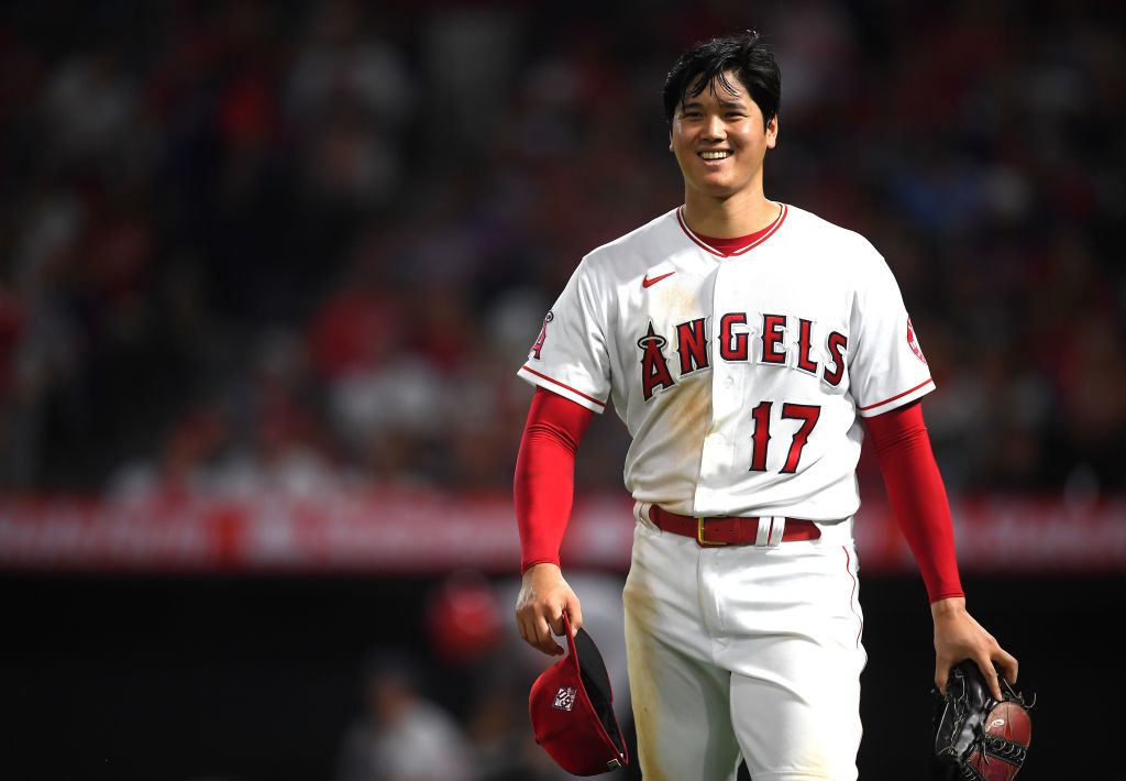 Shohei Ohtani, a baseball player in an Angels jersey with the number 17 on it, holds a baseball cap and glove in hand