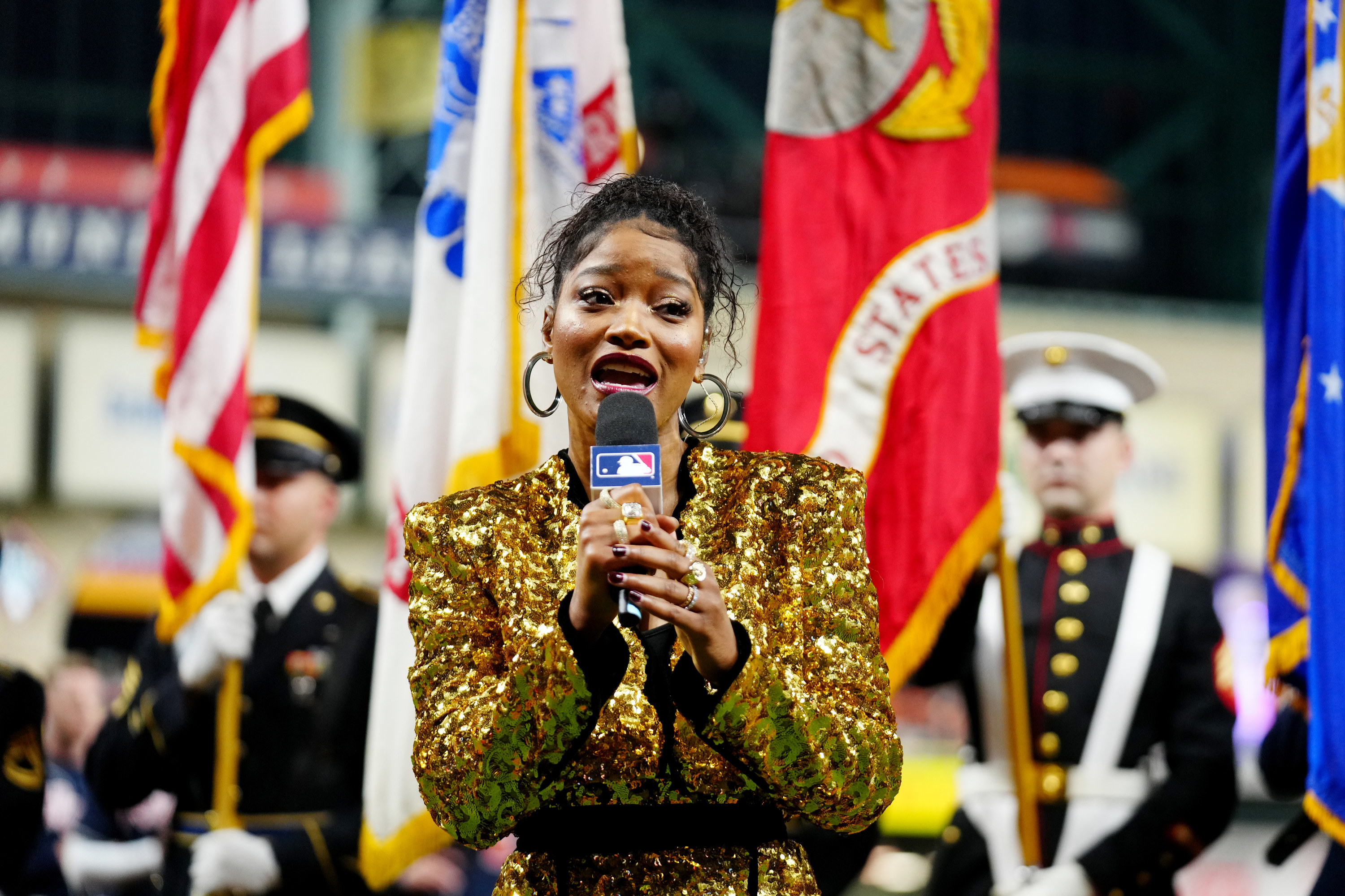 Keke at a microphone singing in front of members of the military in uniform holding flags