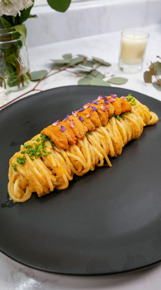 neatly twirled spaghetti with uni sauce on a plate with edible flowers, chives, and uni on top