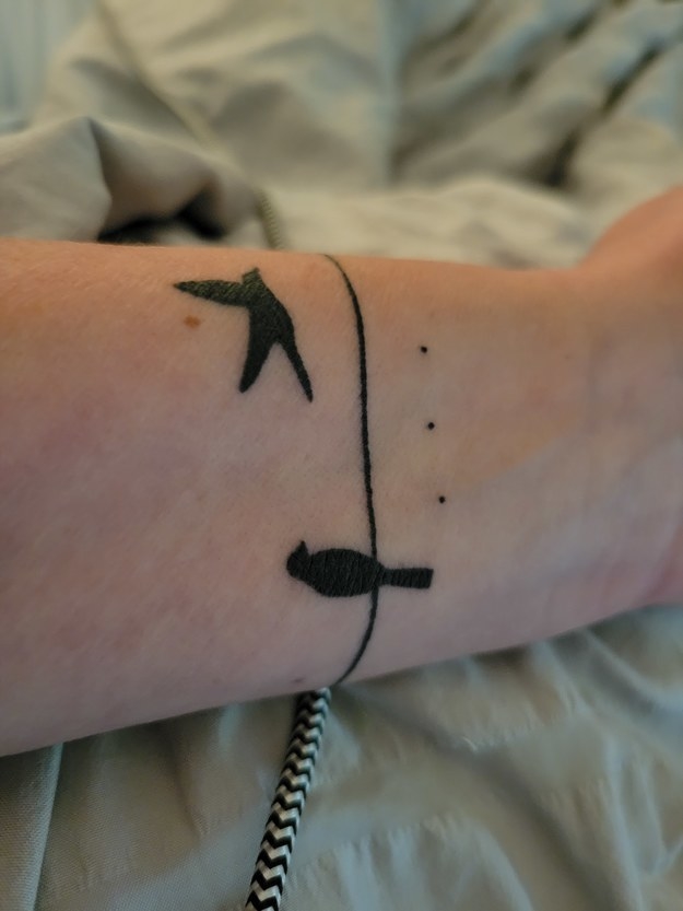 tattoo on arm of bird sitting on wire next to bird flying away with three dots underneath
