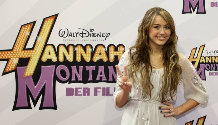 Miley Cyrus smiling in front of a Hannah Montana poster