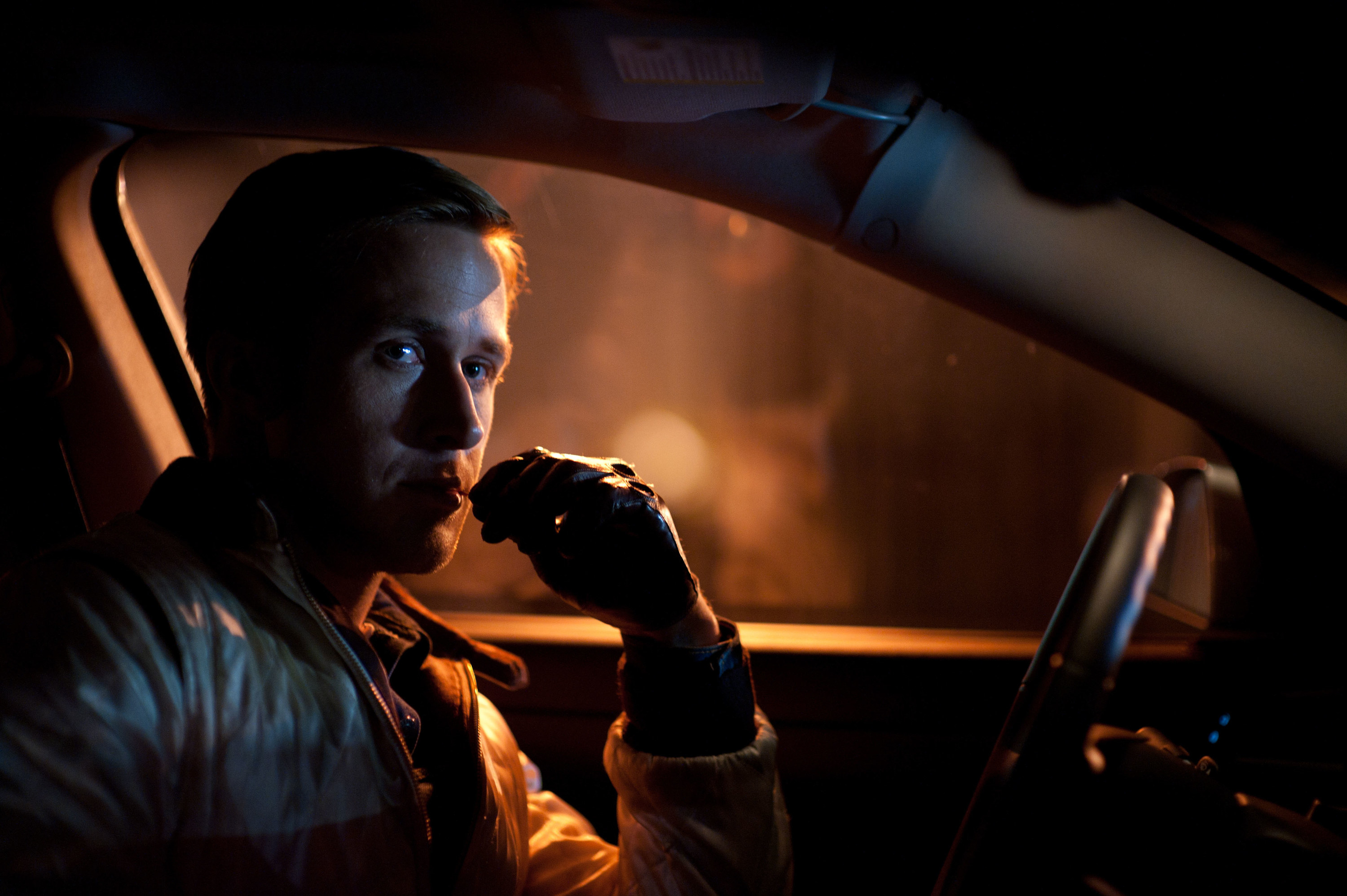 Ryan Gosling sits in a car, holding a toothpick to his mouth with leather gloves