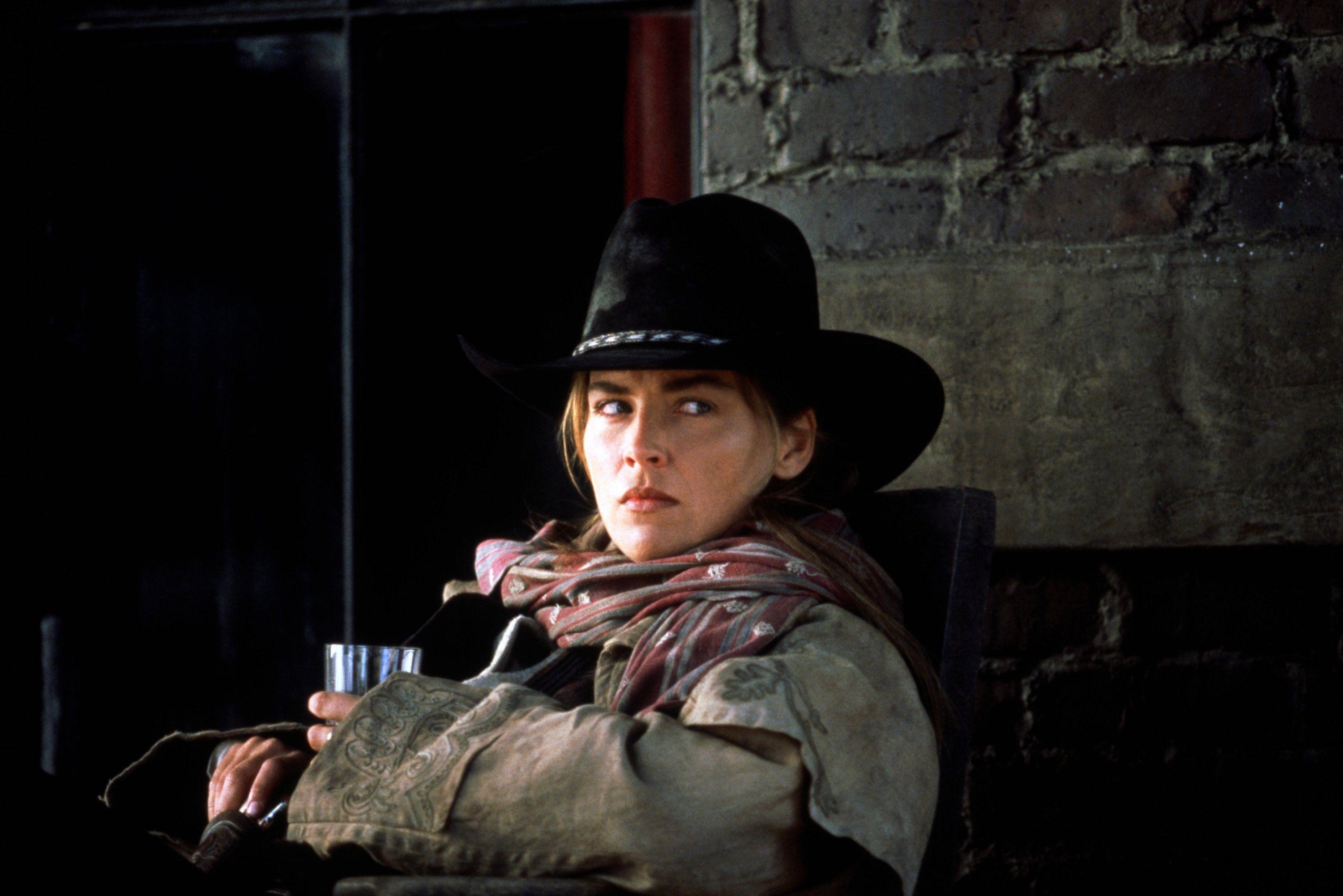 Sharon Stone in a dusty cowboy get-up stares intensely while holding a glass cup and a holstered gun