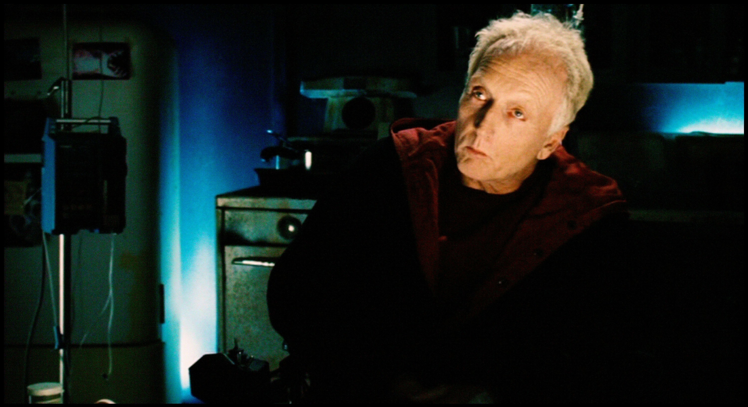 Tobin Bell sits in a dark industrial room in a black-and-red cape
