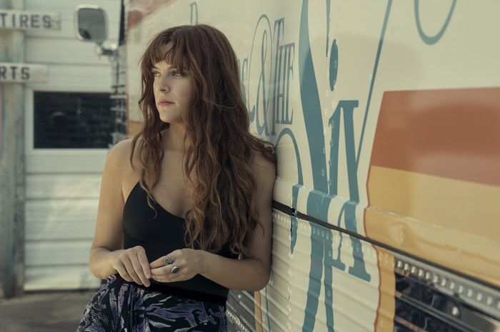 Riley Keough as Daisy Jones stands next to a van bus and looks into the distance