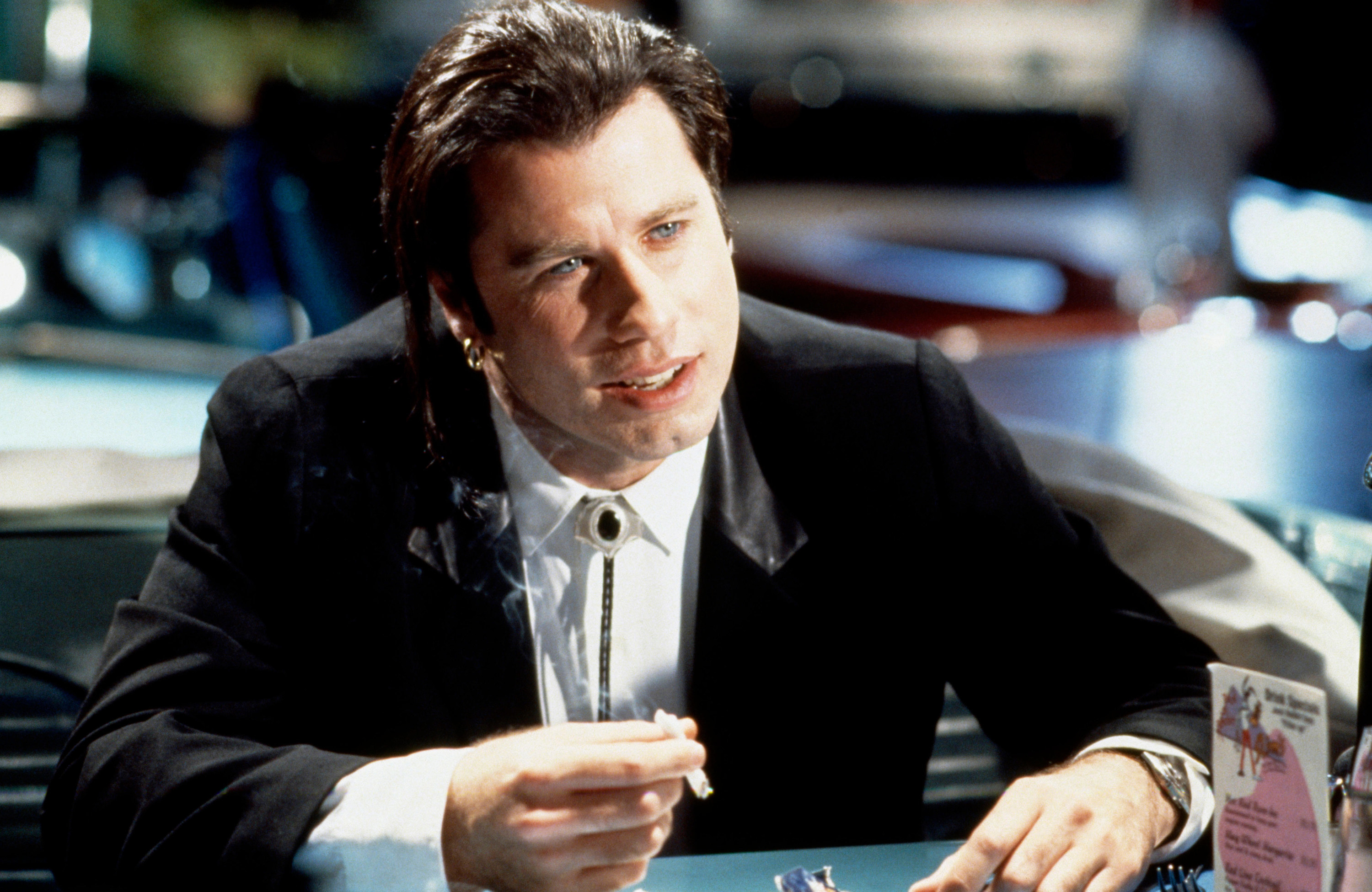 John Travolta in a black suit and bolo tie smokes a cigarette at a theme restaurant