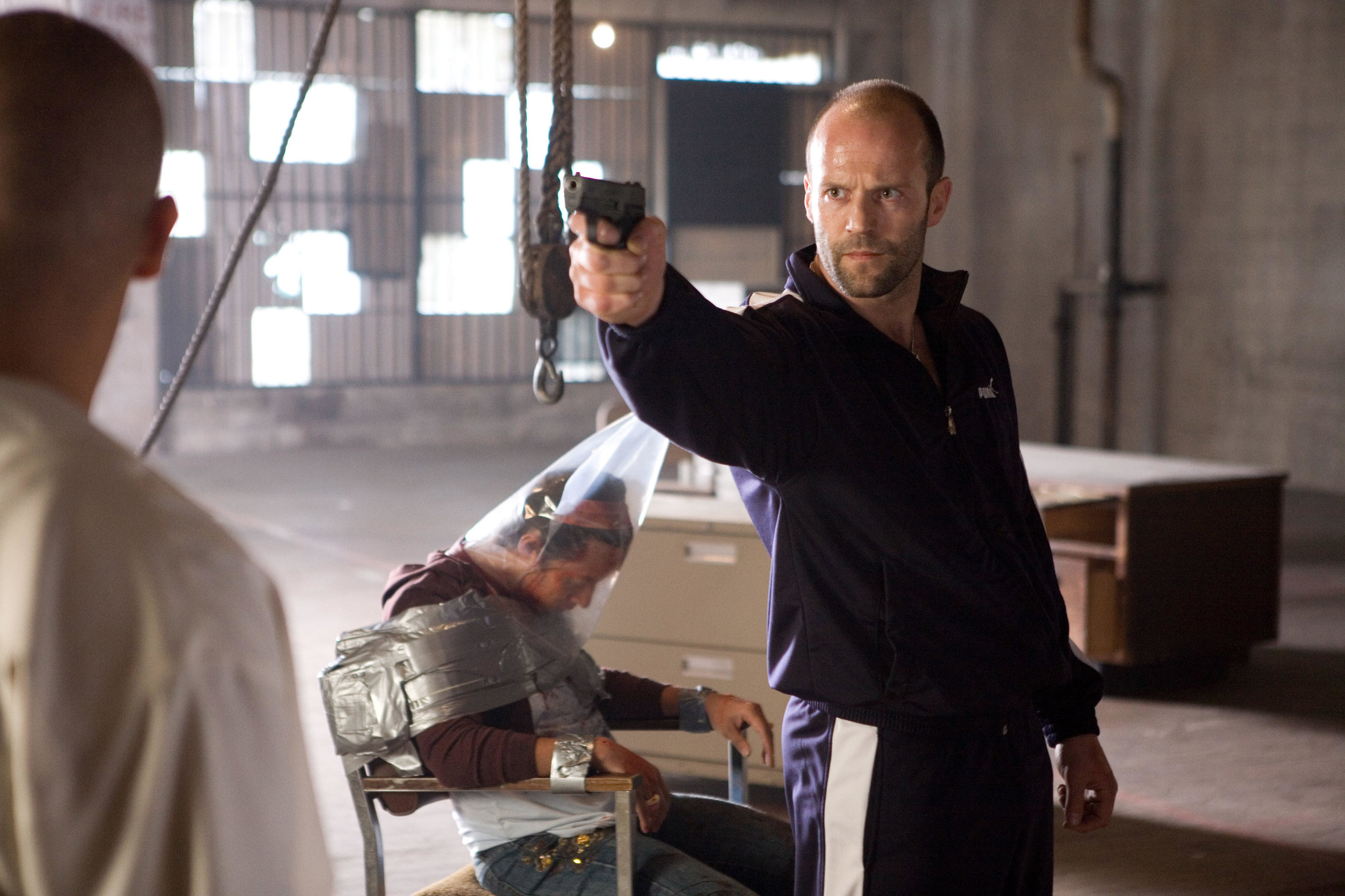 Jason Statham in a track suit holds a gun to a gangster in a warehouse near a dead body