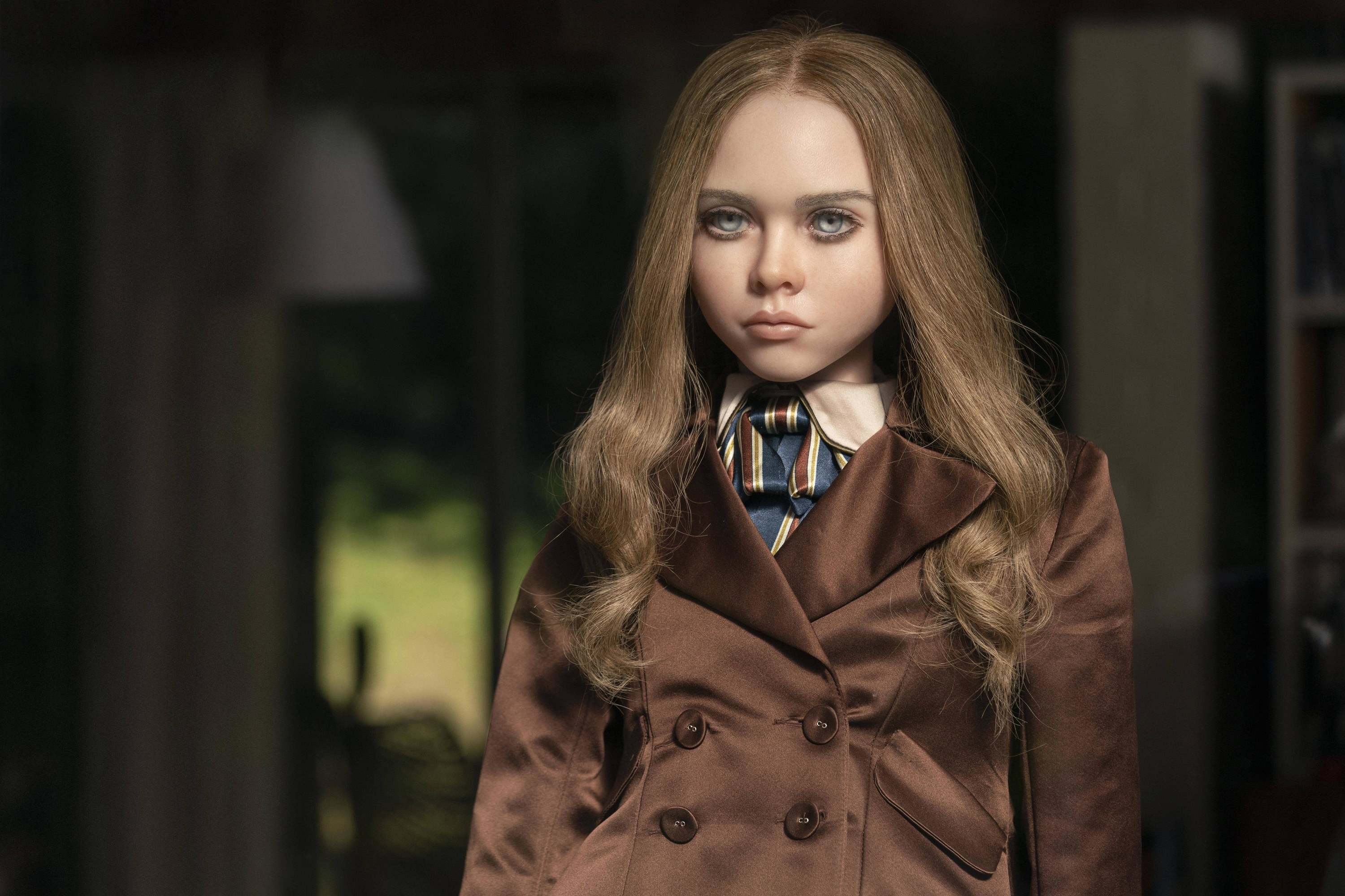 A life-like little girl doll in a brown button-up coat stares ominously outside of a window