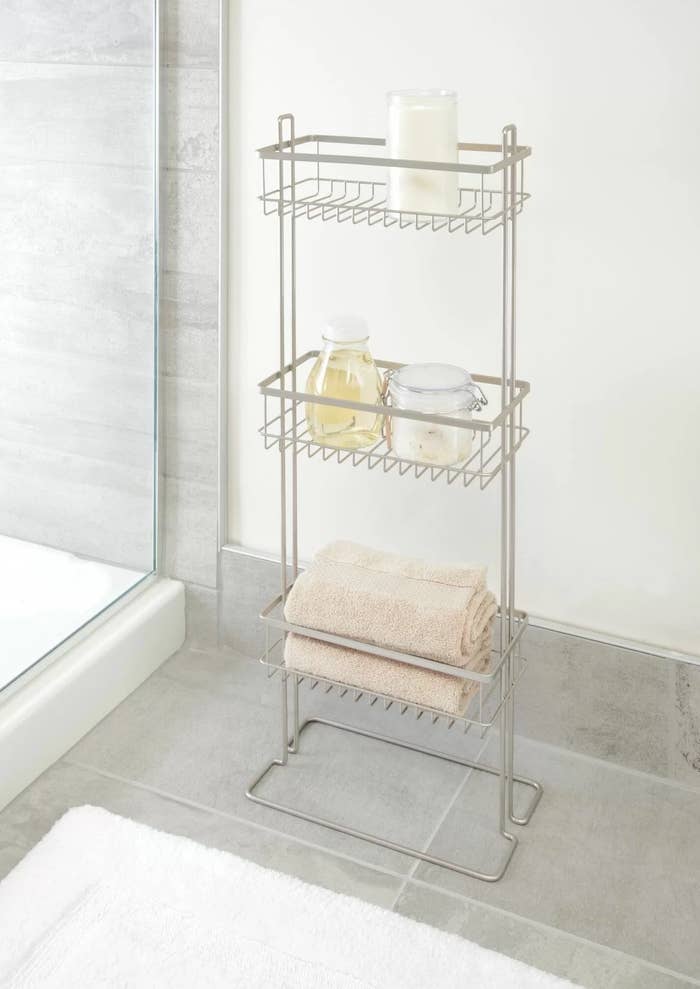 a silver freestanding shower shelf next to a white rug and glass shower door