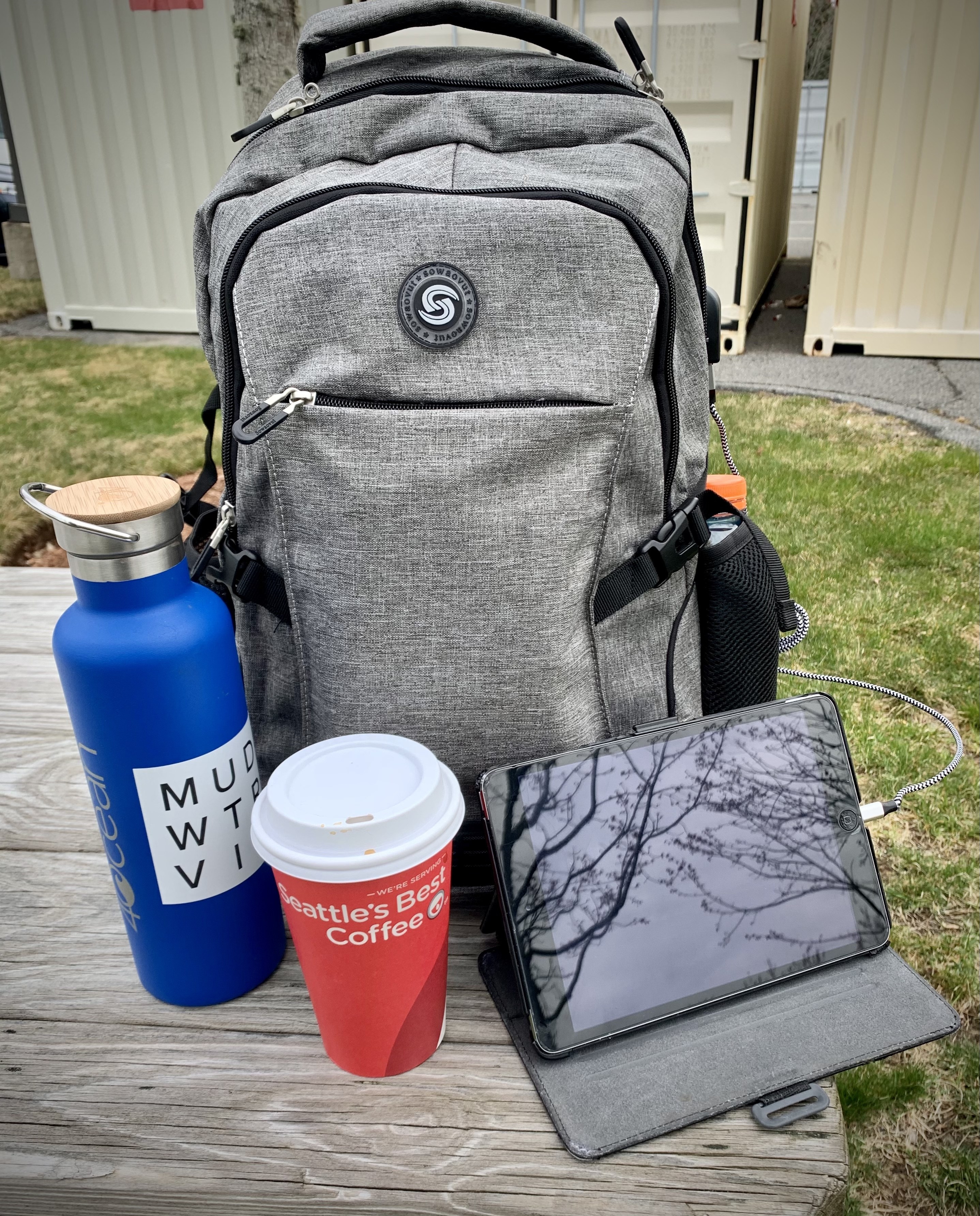 reviewer&#x27;s grey backpack on picnic bench outside charging iPad with water bottle and coffee cup next to it