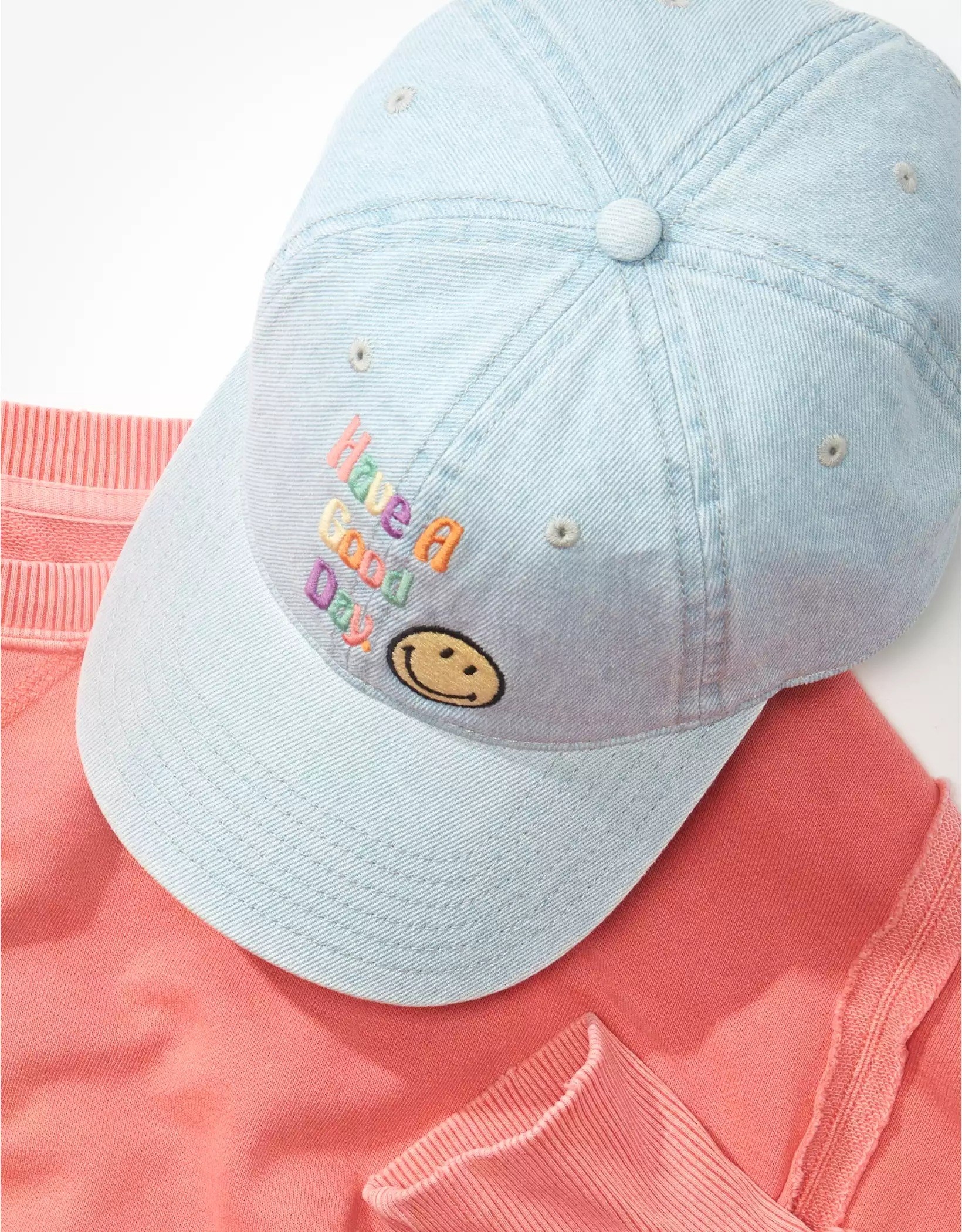 The denim cap with an embroidered smiley face and the words &quot;have a good day&quot; on the front sitting atop a salmon sweatshirt