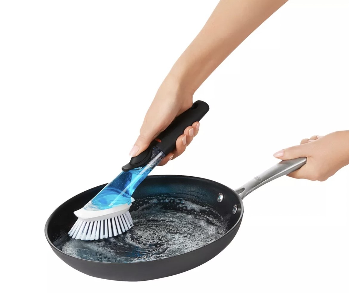 A hand scrubbing a pan with a soap dispenser brush