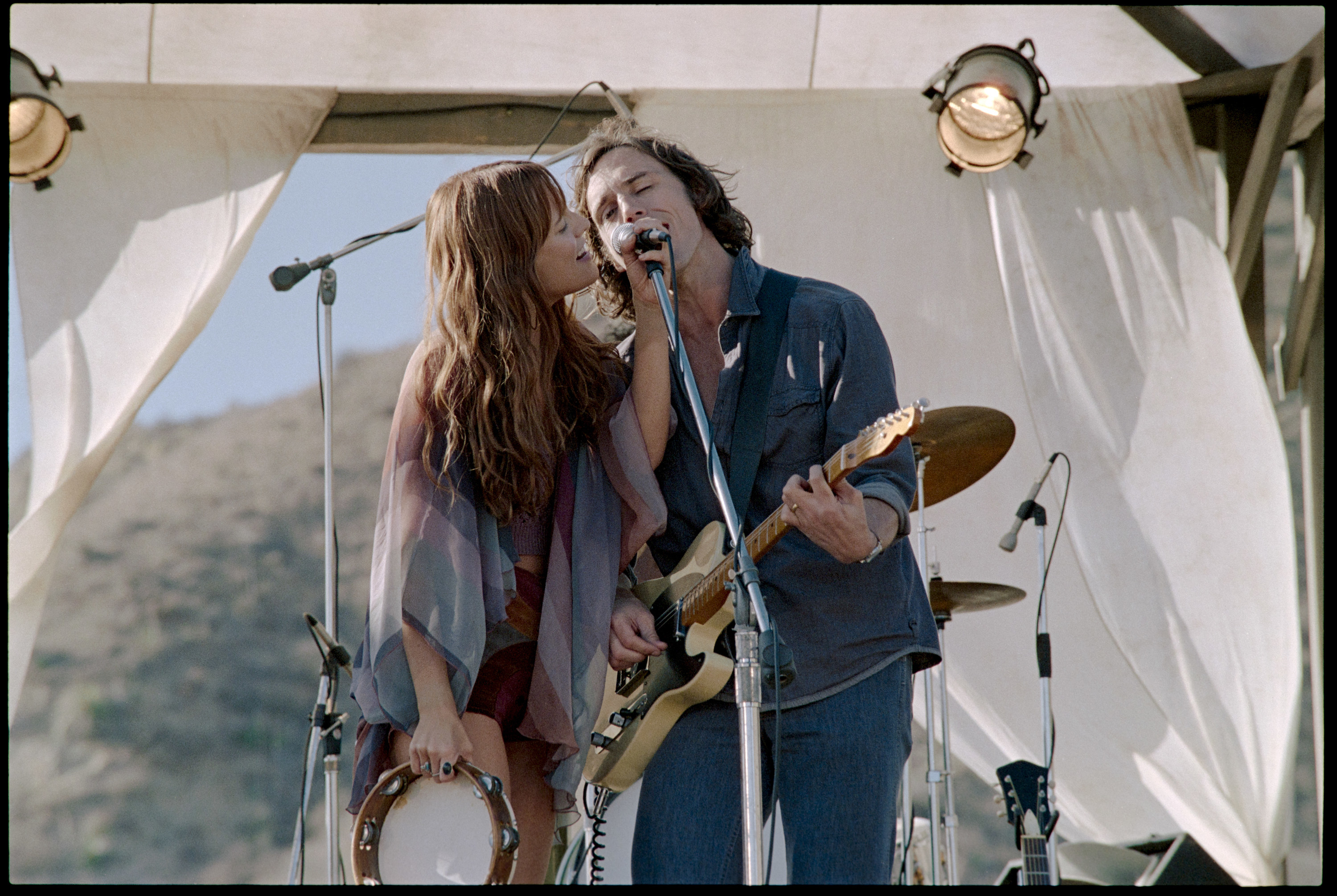 Daisy and Billy sing into the same microphone as Billy plays a guitar and Daisy holds a tambourine