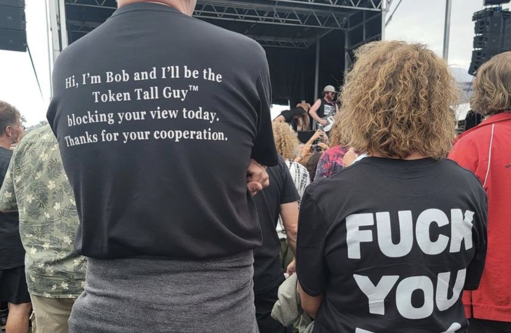 Tall guy with back of T-shirt saying &quot;Hi, I&#x27;m Bob and I&#x27;ll be the Token Tall Guy blocking your view today, thanks for your cooperation,&quot; and the woman with &quot;Fuck you Bob&quot; on the back of her shirt