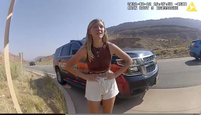 A still from a police body camera shows a young woman with her hadns on her hips, standing by a police truck on the side of a road in an arid, hilly landscape