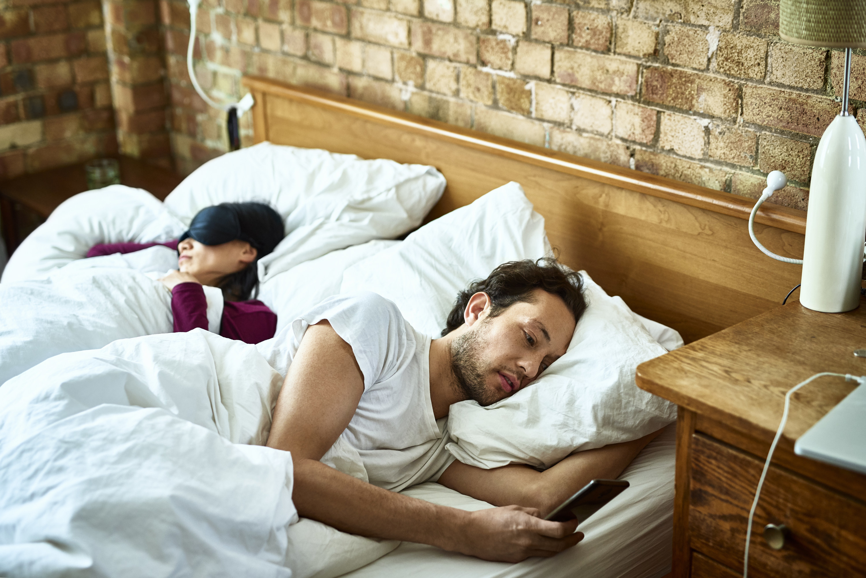 A man looking at his phone and turned away from his sleeping partner in bed