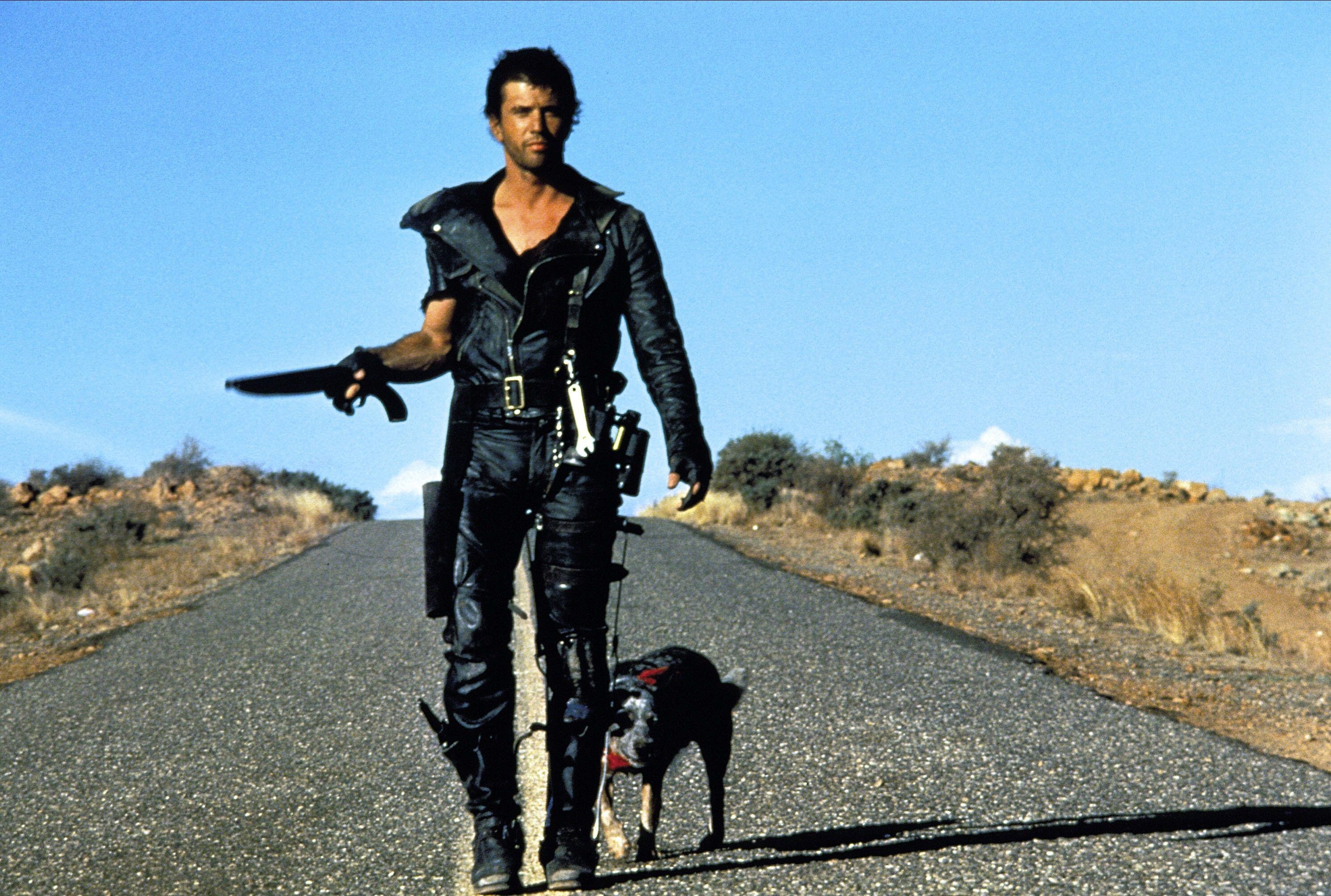 A young Mel Gibson in a modified police officer outfit wanders down an empty road with a dog in tow
