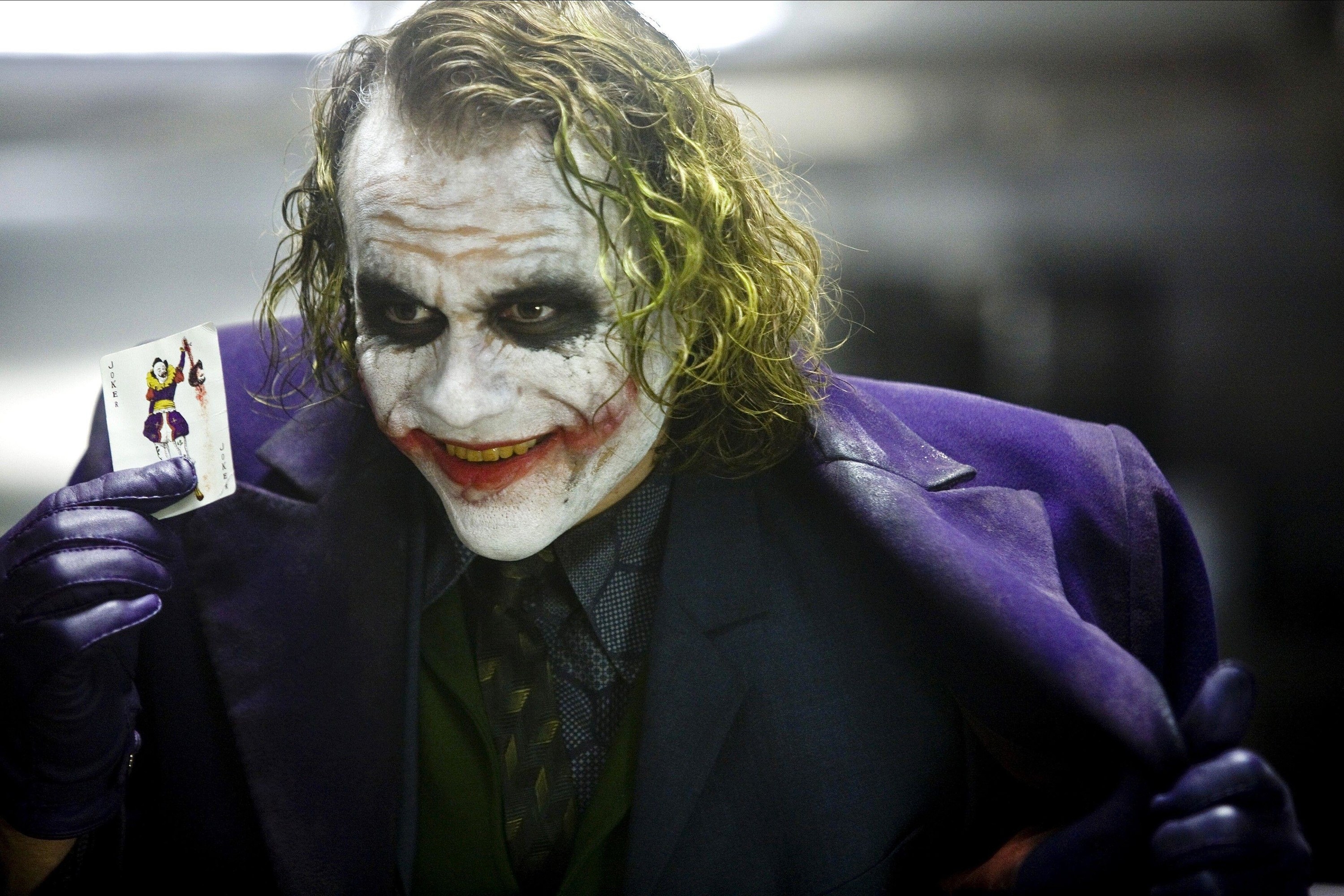 Heath Ledger as the Joker holds up a playing card ominously while holding open his purple jacket