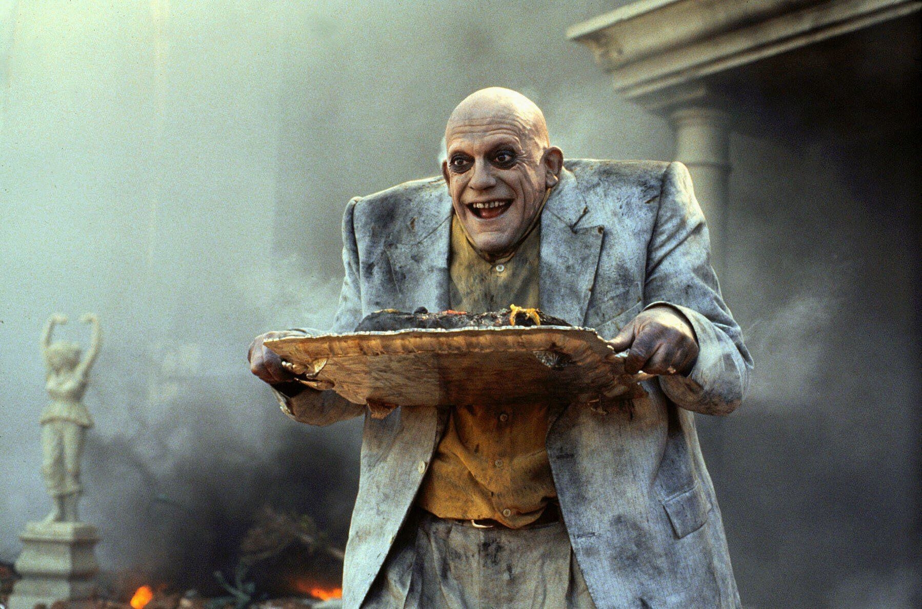 A bald man in a suit, charred from an explosion, stands in the burning wreckage holding a silver platter