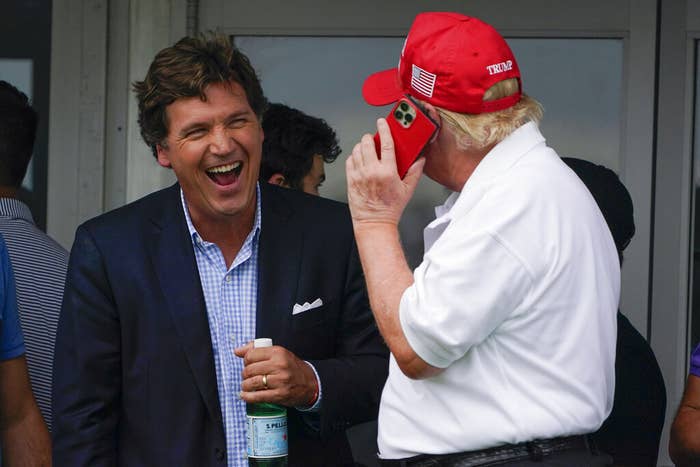 A man in a suit and a light blue plaid shirt laughs and holds a bottle of sparkling water beside Donald Trump, who has his back to the camera and is wearing a red Make America Great Again hat and has a red iPhone up to his ear