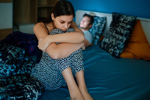 Sad girlfriend sitting on bed edge, holding her legs after fight, her partner is lying in bed in background, his face is lit by smart phone