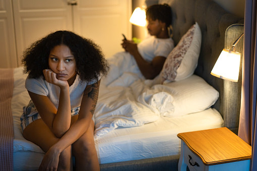 A frustrated woman sitting on the edge of the bed, while her unconcerned boyfriend in scrolling through his smart phone, lying behind her