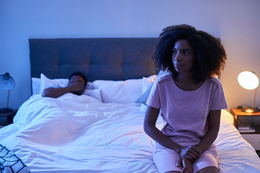 woman sitting at the end of the bed, looking like she&#x27;s thinking about something, while the man lays in bed rubbing his face