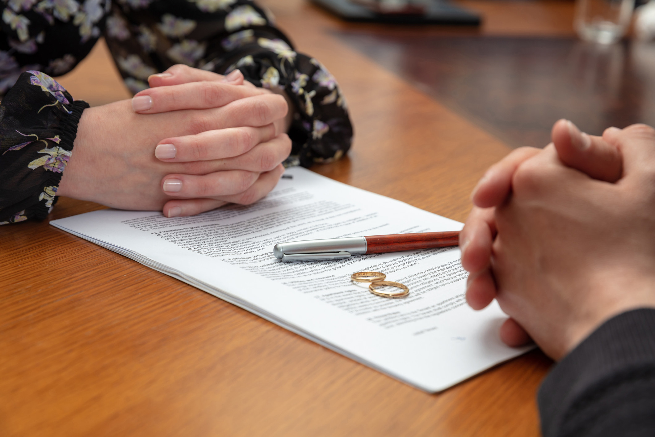 Two people&#x27;s hands next to papers with wedding rings and a pen on top