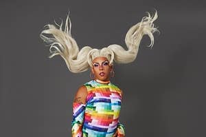 Jax from Season 15 of Ru Paul's drag race boldly strikes a pose with her feet apart and blonde pigtails flying above her. The queen is wearing a rainbow prism outfit with thigh high boots, a matching unitard, and fingerless gloves. 