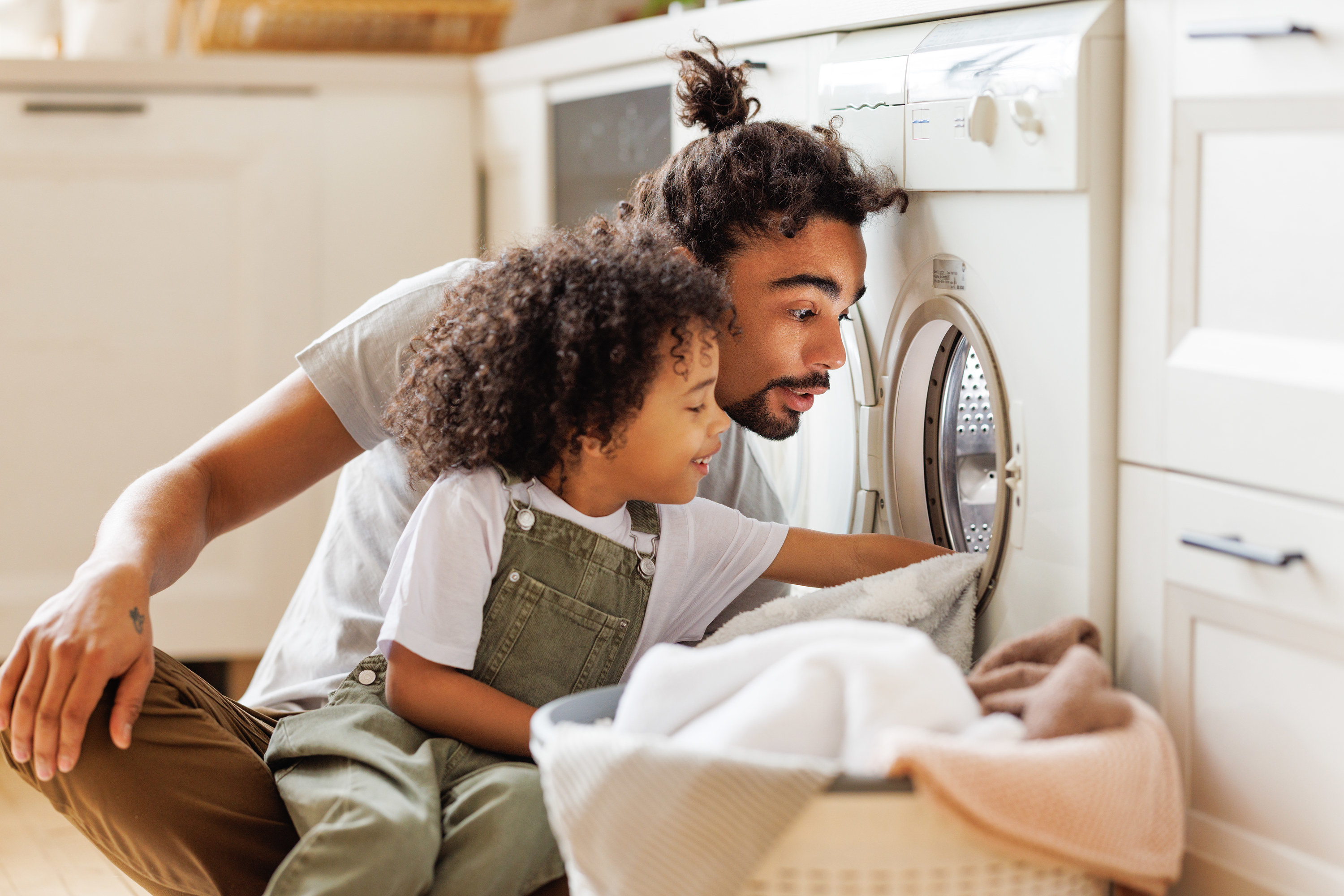 Cheerful black kid boy sitting on dad&#x27;s lap and helping father at linen in basket while doing laundry near washing machine in flight kitchen in weekend at home