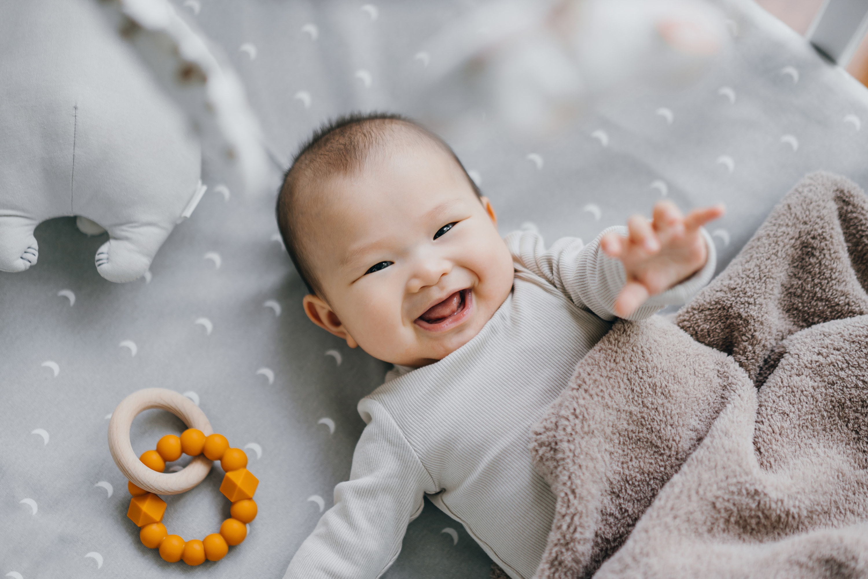 Portrait of a lovely Asian baby girl smiling sweetly while lying on the crib. She is raising her hand trying to touch the cot mobile. Baby&#x27;s growth and development concept
