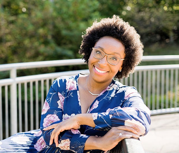 A woman with brown natural curls wearing a navy floral shirt and sitting outdoors in the sun