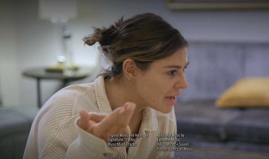 Raquel sitting across from Lala in bed, holding her hand in the air