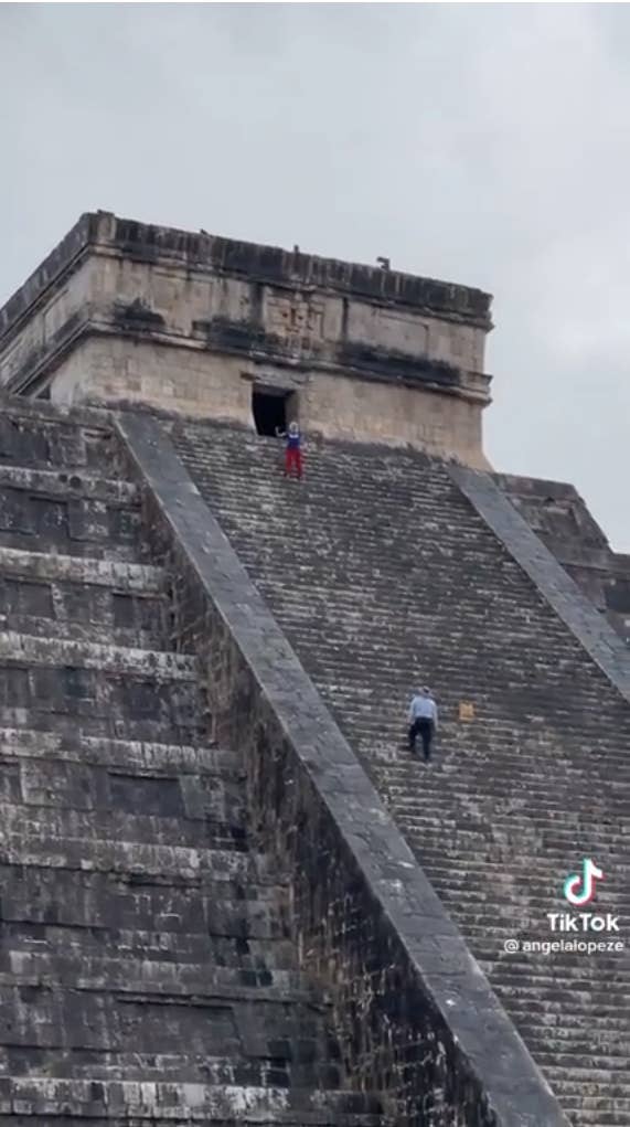 A screenshot shows a woman posing for the camera at the top of a pyramid while a worker climbs up the pyramid to stop her