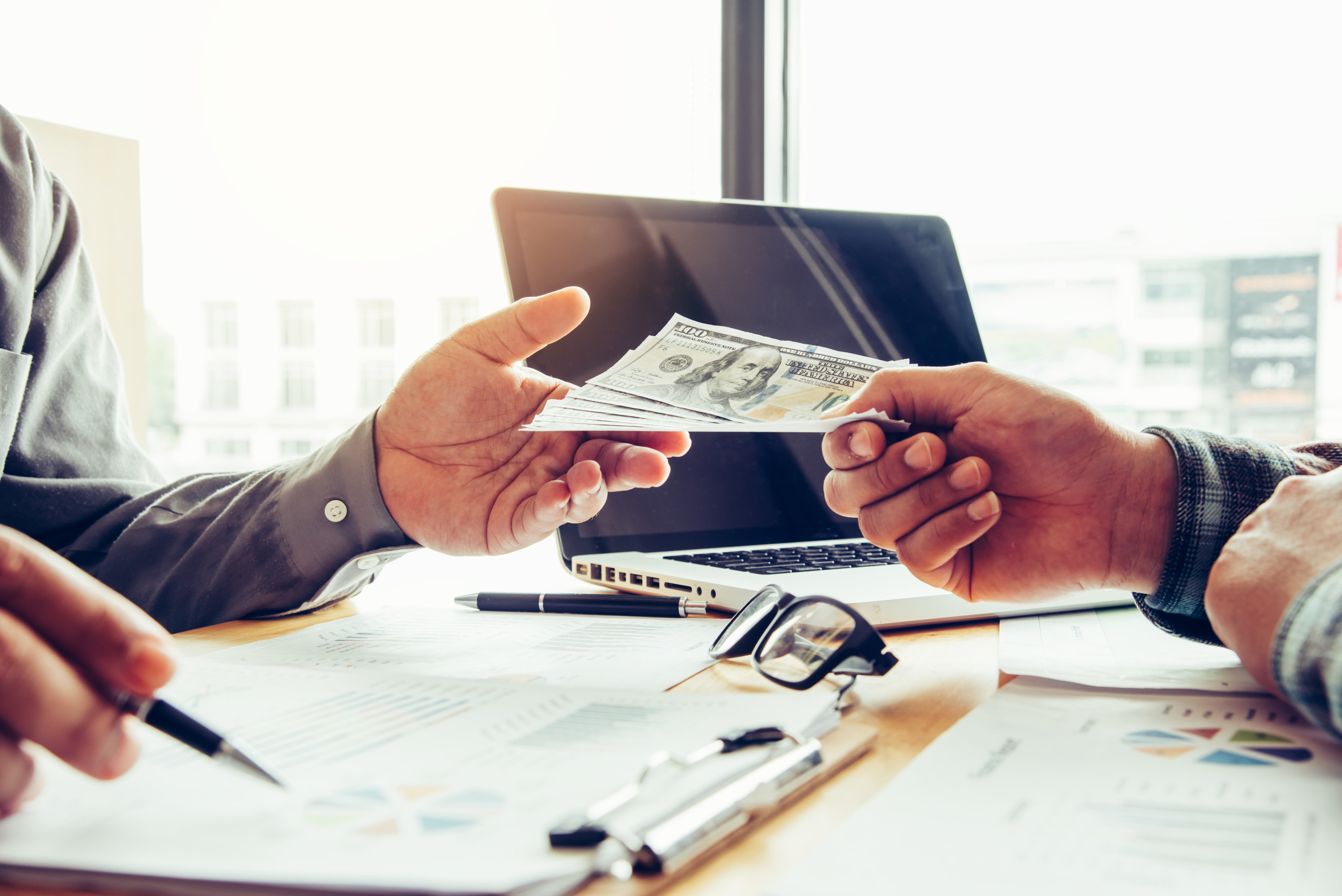 Two men passing cash across a table over a laptop