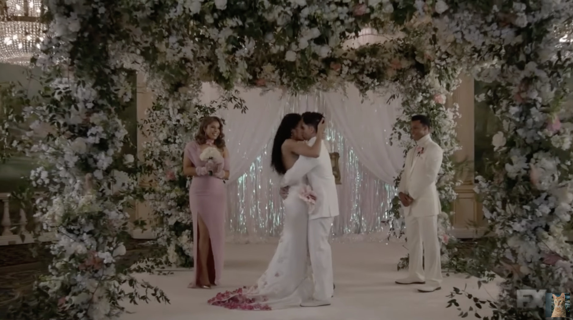 A couple wearing white stands at the altar embraced in a passionate kiss, under a huge flowering arch, as a bridesmaid in pink and a groomsman in a cream suit stand beside them