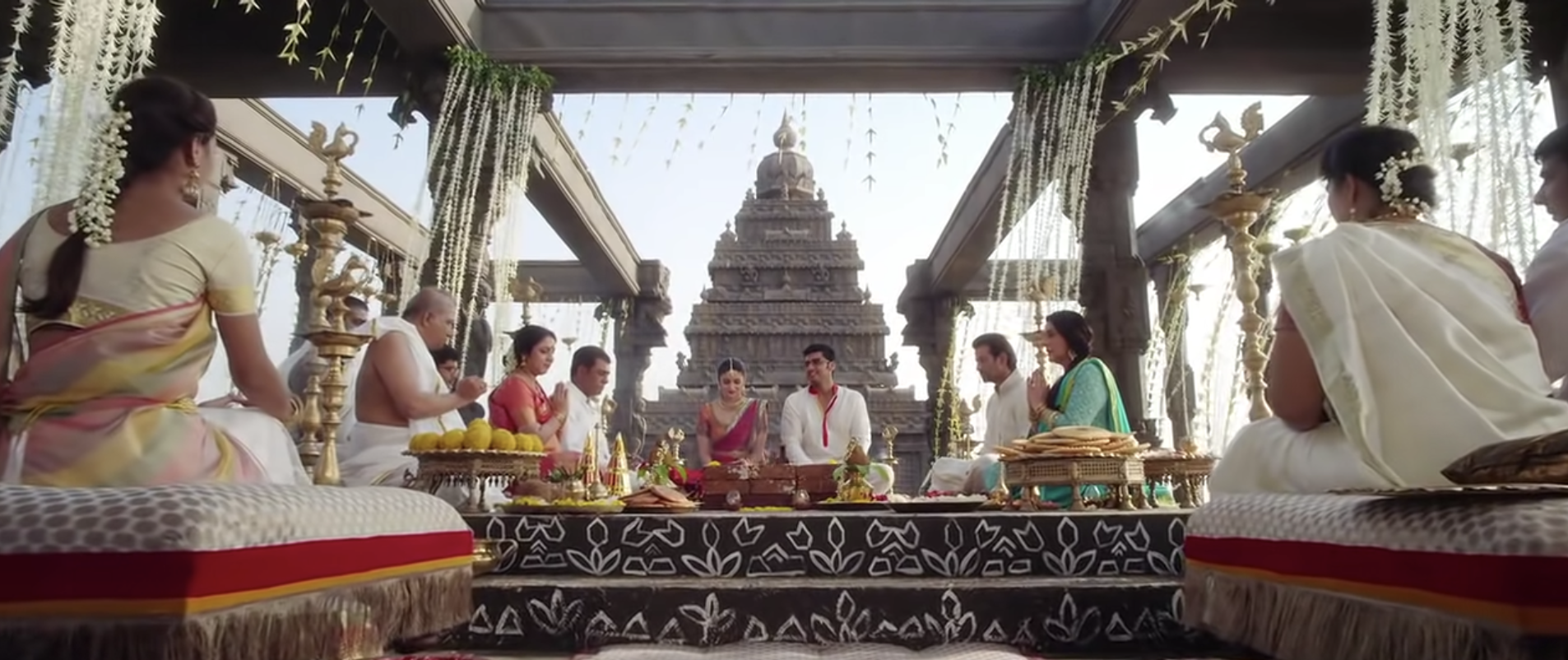 A couple sits cross legged at an altar, framed by family members also sitting, with a temple behind them and more guests sitting and watching