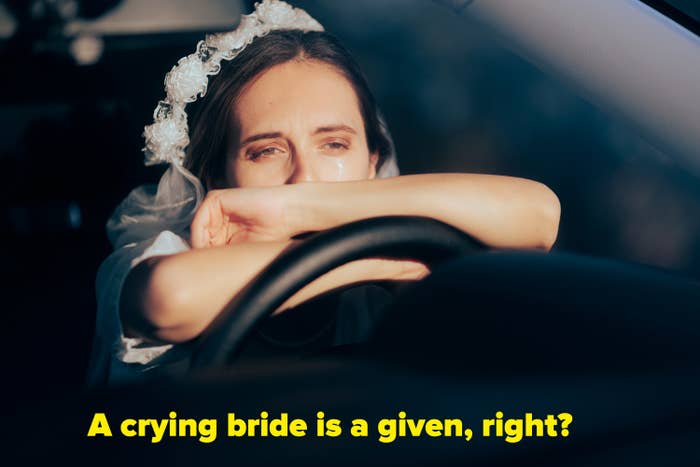 A crying bride wearing a wedding veil leaning with both arms on the steering wheel of a car.