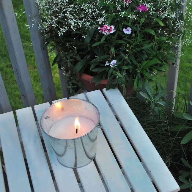 a lit citronella candle on a wooden outdoor table next to a plant